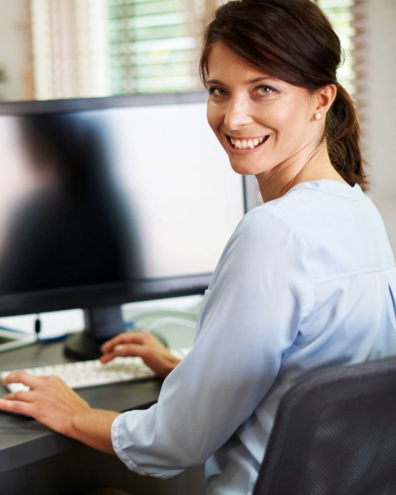 Funcionária sorrindo no computador do escritório, com uma colega ao fundo