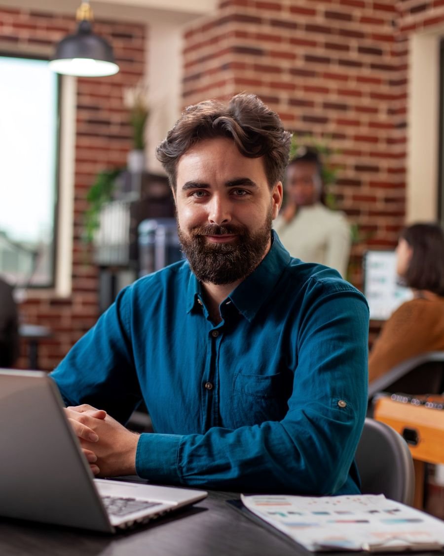 Empresário sorrindo diante do notebook, com a equipe trabalhando nos computadores ao fundo